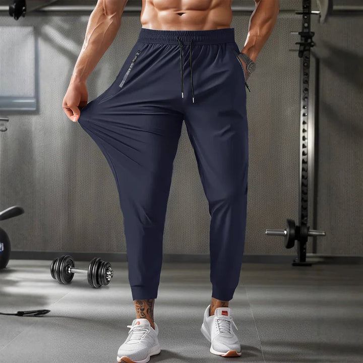 Muscular man stretching navy blue jogger pants in modern gym with dumbbells and workout equipment