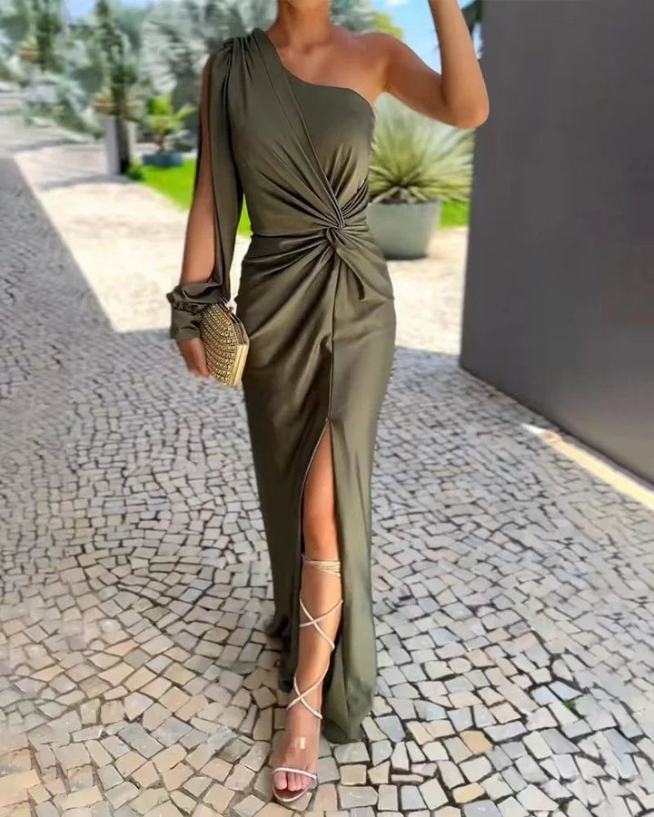 Woman wearing olive green one-shoulder evening dress with front slit and holding clutch on stone tile path