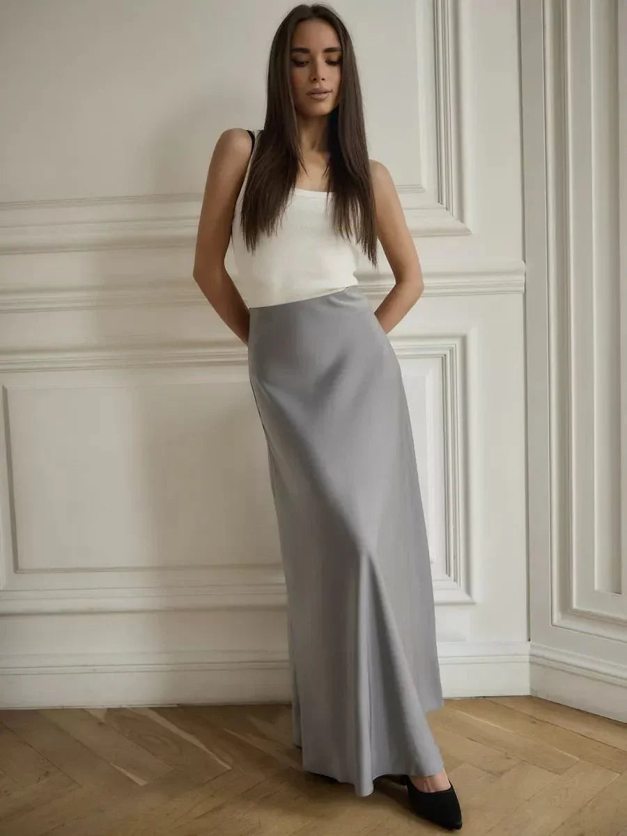 Woman wearing a white sleeveless top and long gray skirt standing in a minimalist room with wood floor