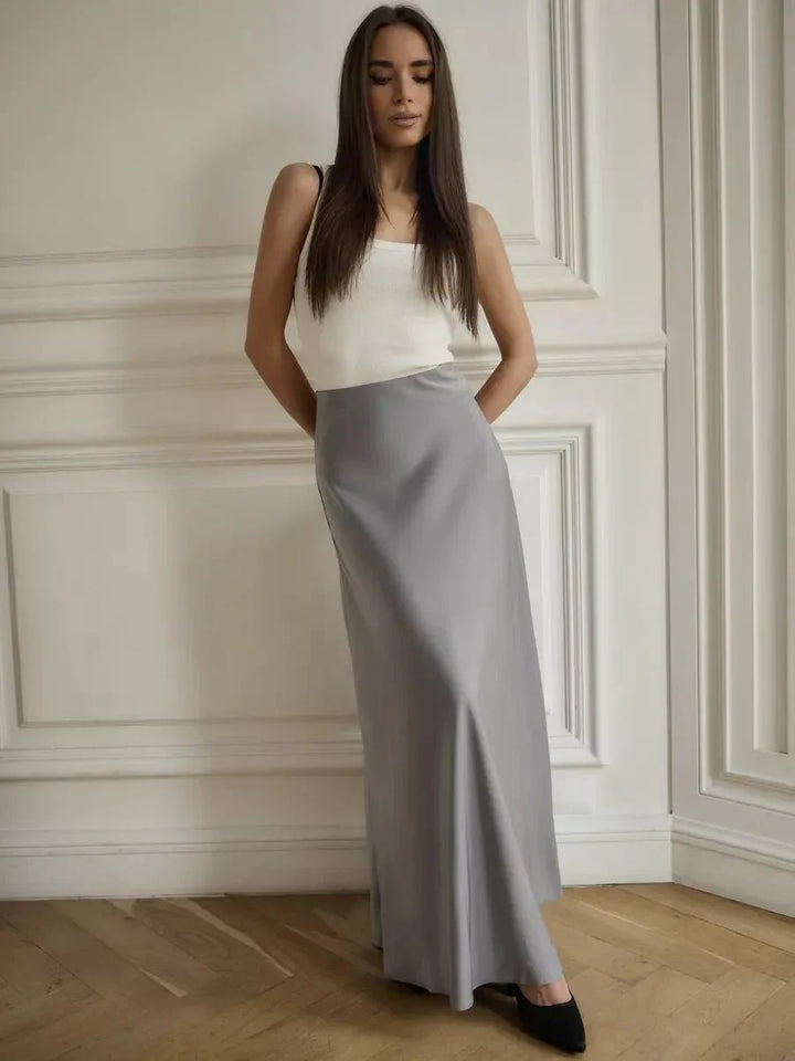 Woman wearing a white sleeveless top and long gray skirt standing in a minimalist room with wood floor