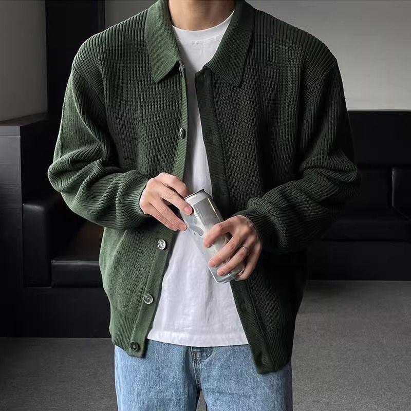 Man wearing dark green knitted cardigan over white t-shirt and blue jeans holding a drink can indoors