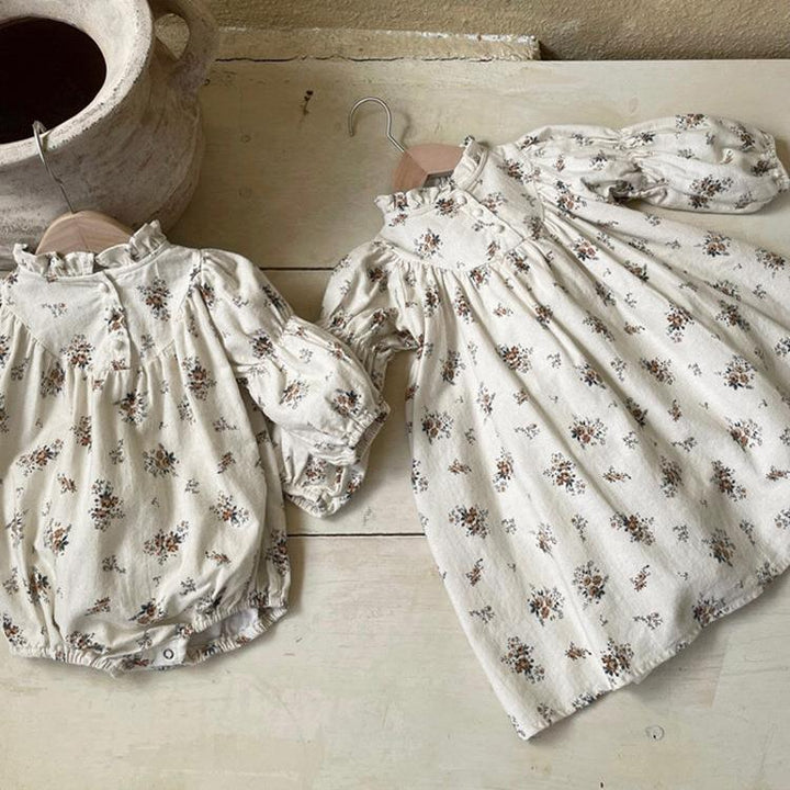 Two cream-colored baby outfits with floral prints and puffed sleeves on wooden hangers on a rustic wooden surface