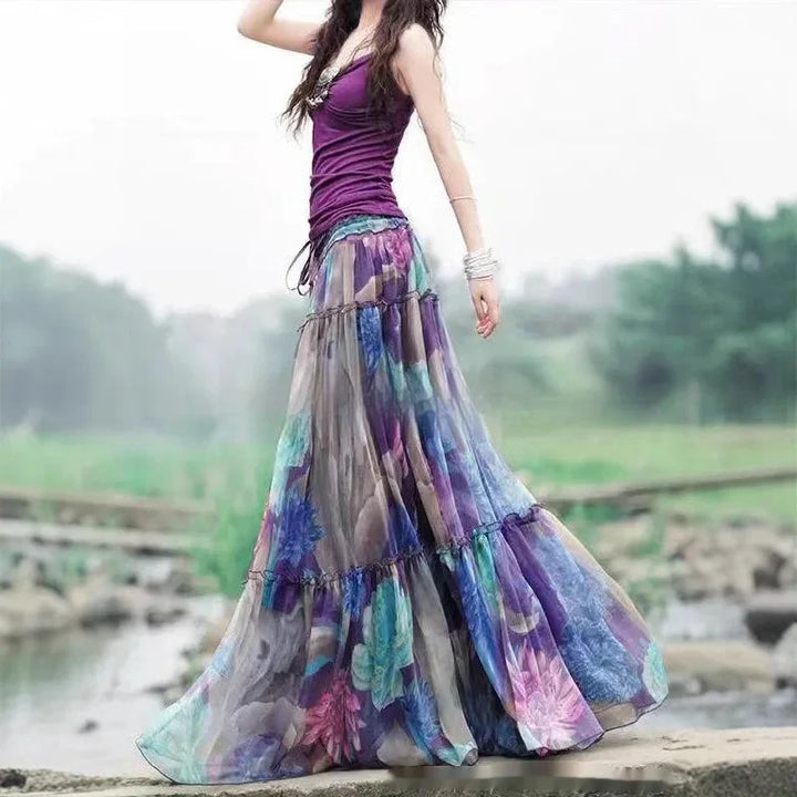 Woman in purple tank top and flowing colorful floral maxi skirt outdoors by water and stone path