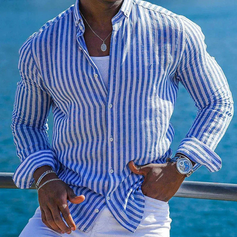 Man wearing blue and white striped shirt, white pants, silver watch, and necklace by ocean railing