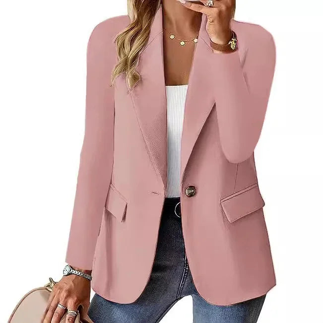 Woman wearing a pink blazer over white top and blue jeans, accessorized with gold jewelry and holding a round beige bag