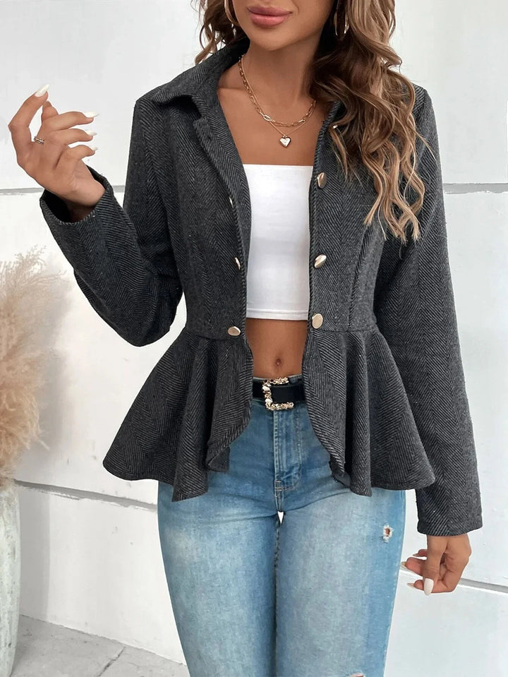 Woman wearing a dark gray herringbone peplum blazer with silver buttons over white crop top and blue high-waist jeans