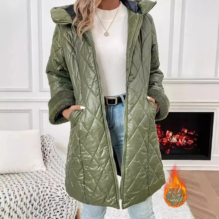 Woman wearing quilted olive green winter coat with fleece cuffs, white sweater, and jeans near fireplace