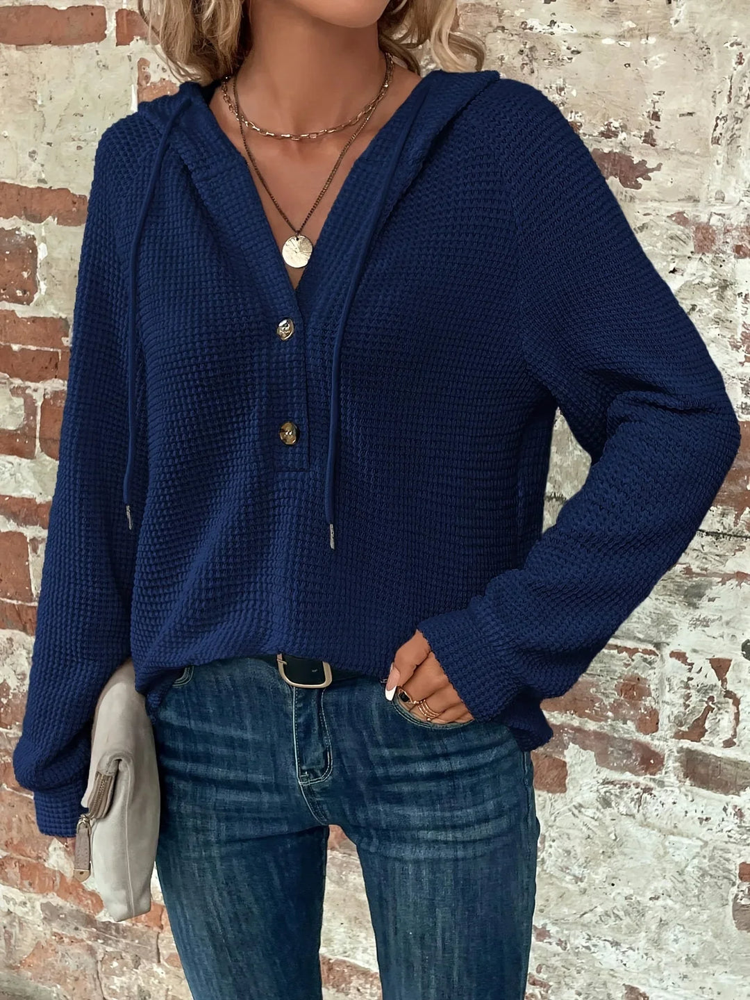 Woman wearing navy blue waffle knit hoodie with gold buttons and blue jeans against brick wall