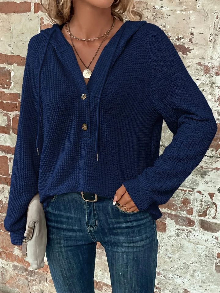 Woman wearing navy blue waffle knit hoodie with gold buttons and blue jeans against brick wall