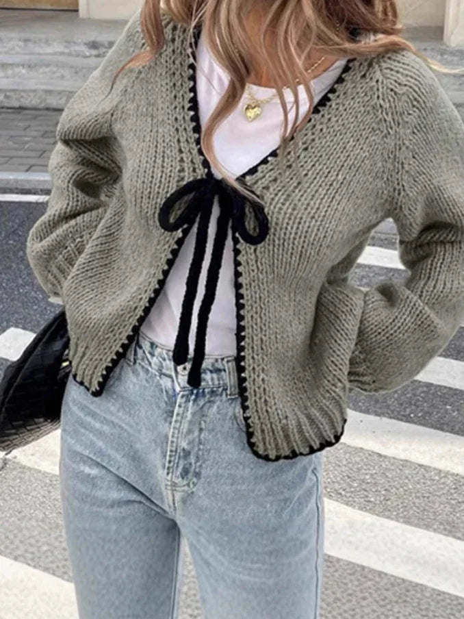 Woman wearing gray knitted cardigan with black trim and tie, white top, light blue jeans, and gold heart necklace