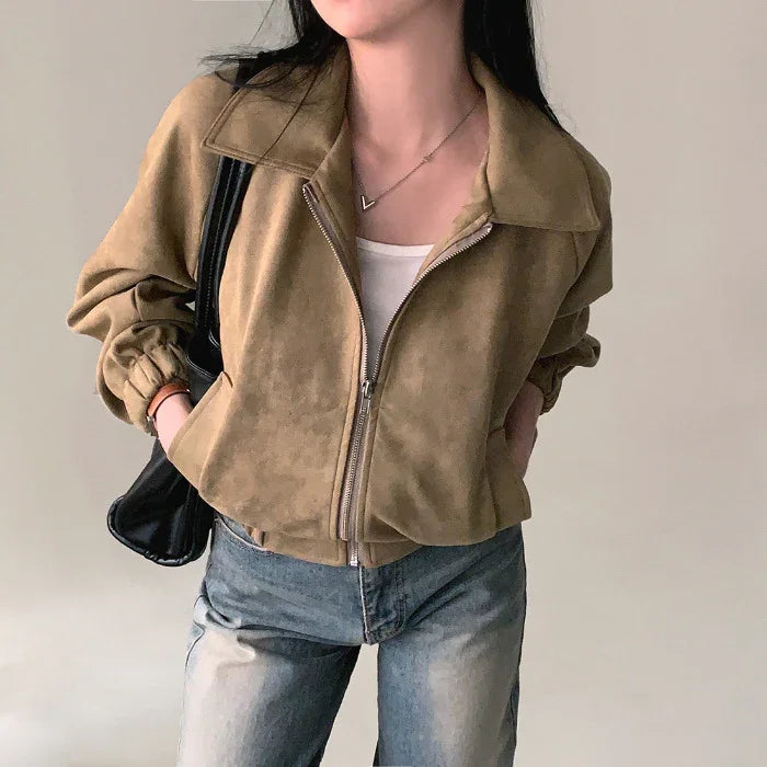 Woman wearing tan suede zip jacket with elastic cuffs, white top, blue jeans, and black shoulder bag