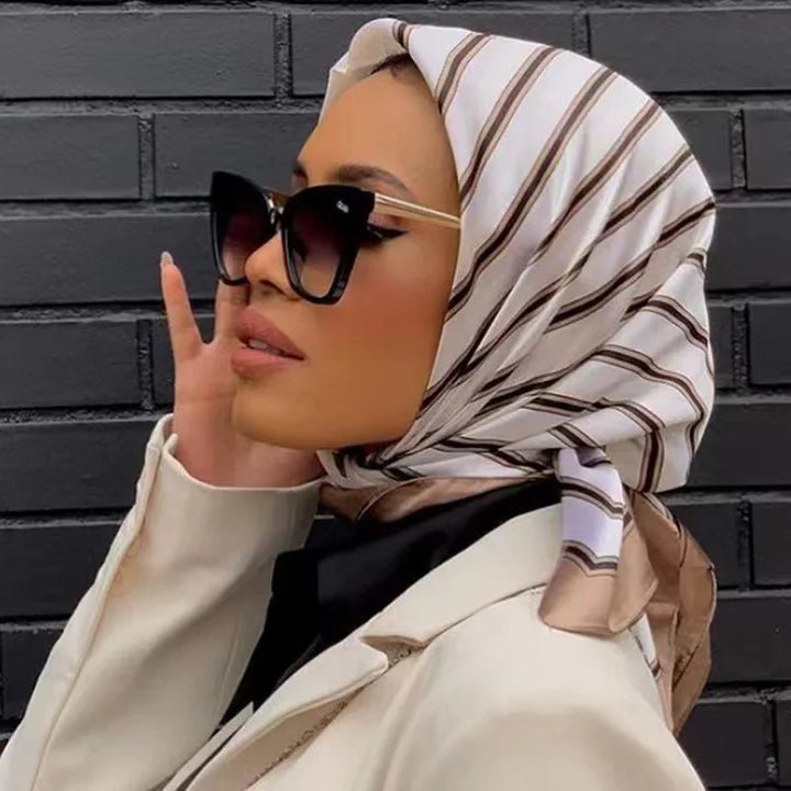 Woman wearing a striped beige and white hijab, oversized black sunglasses, and a cream blazer against a black brick wall