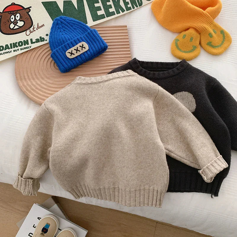 Cozy beige and black children's sweaters on bed with blue knit hat and yellow smiley face socks