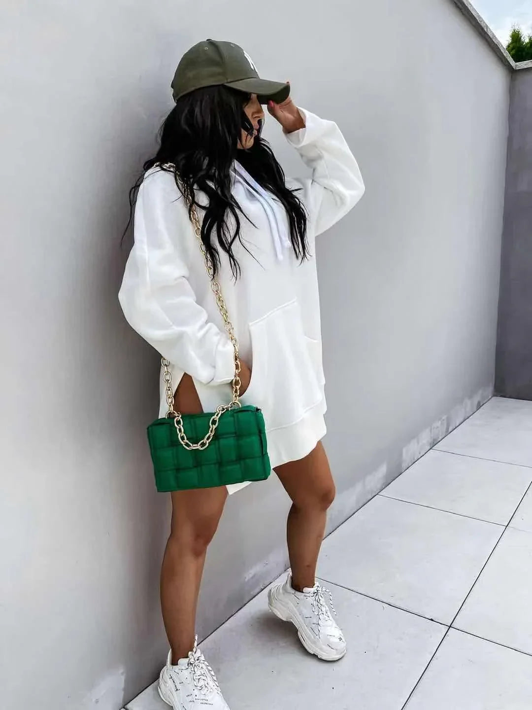 Woman in white oversized hoodie dress, green quilted chain bag, white sneakers, and olive cap leaning on gray wall