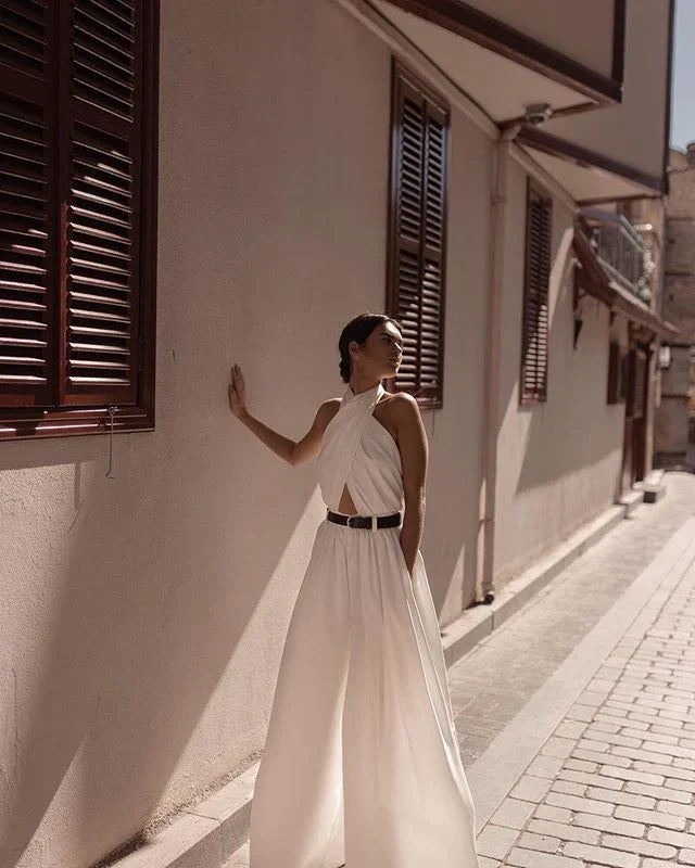 Woman in stylish white halter jumpsuit with black belt standing against sunlit beige wall with wooden windows