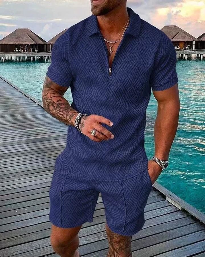 Man wearing navy blue textured short-sleeve shirt and matching shorts standing on wooden pier by turquoise water and overwater bungalows