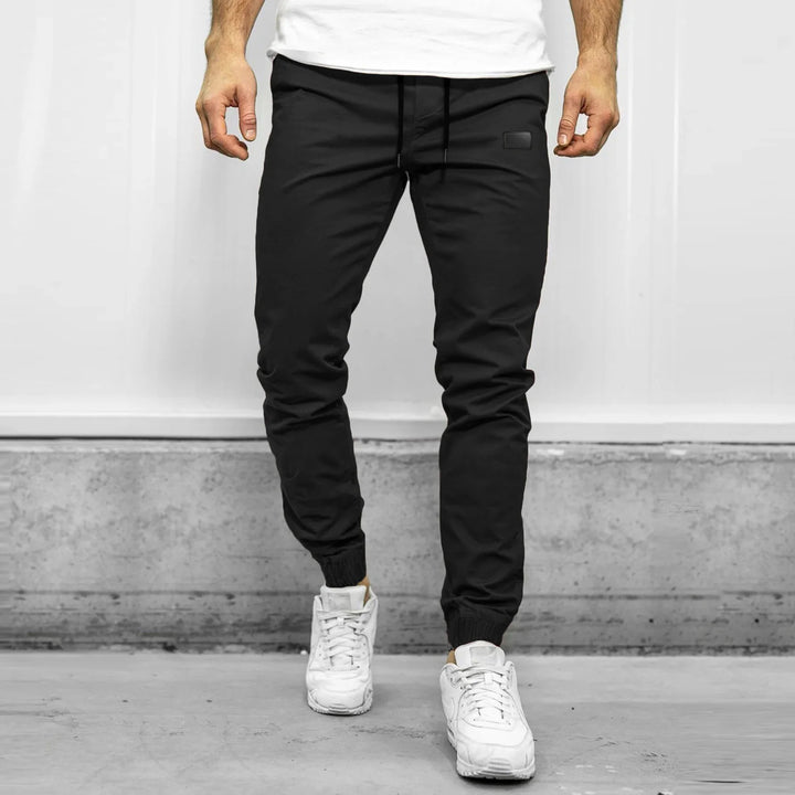 Man wearing black jogger pants and white sneakers standing on concrete floor with white wall background