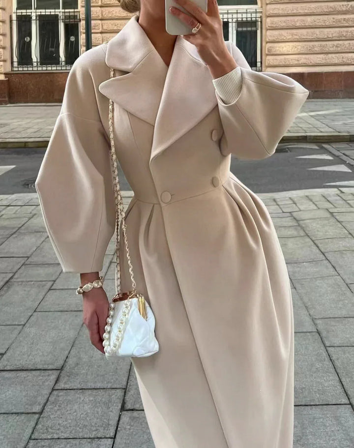 Woman wearing beige double-breasted coat with wide lapels and voluminous sleeves, holding a pearl-accented white handbag on city sidewalk