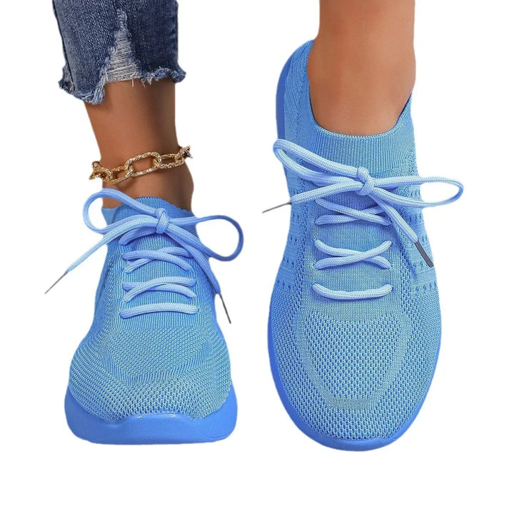 Close-up of feet wearing light blue knit sneakers with matching laces and a gold chain anklet, paired with ripped denim jeans