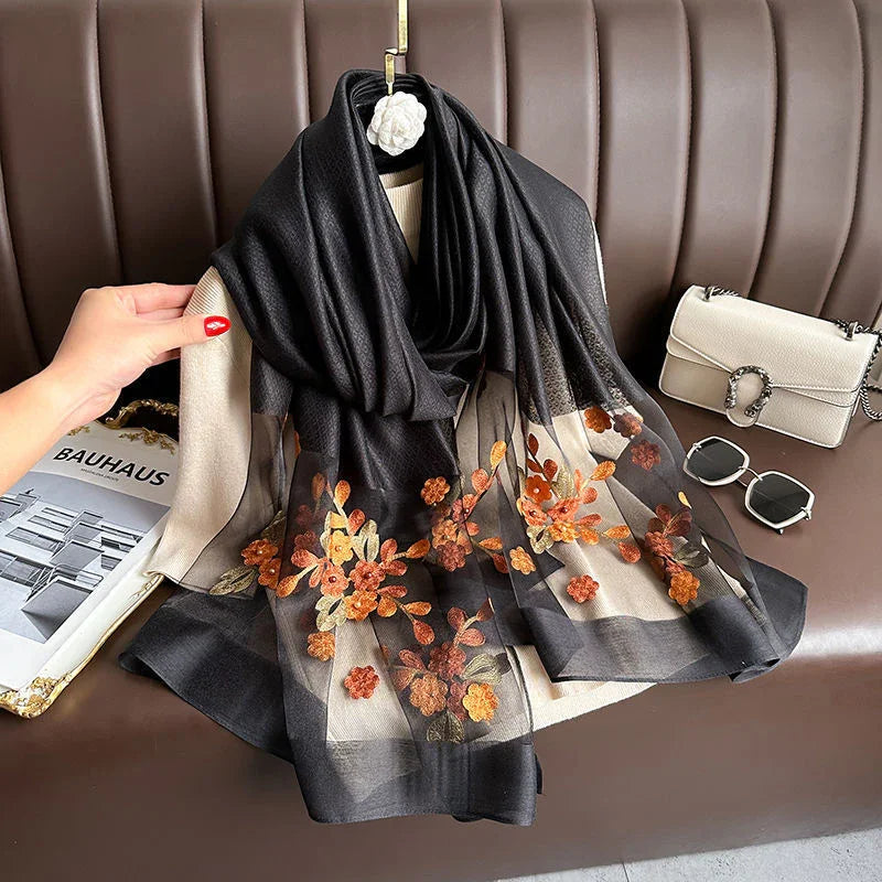 Black semi-transparent scarf with embroidered orange and yellow flowers displayed on beige fabric with white handbag and sunglasses