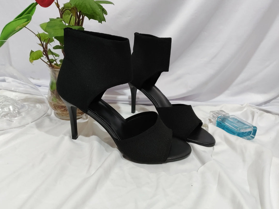 Black high heel open-toe ankle boots on white fabric backdrop with plant and blue perfume bottle