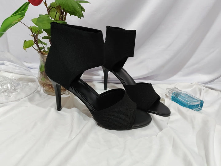 Black high heel open-toe ankle boots on white fabric backdrop with plant and blue perfume bottle