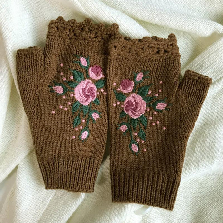 Brown knitted fingerless gloves with pink rose embroidery and green leaves on a white fabric background