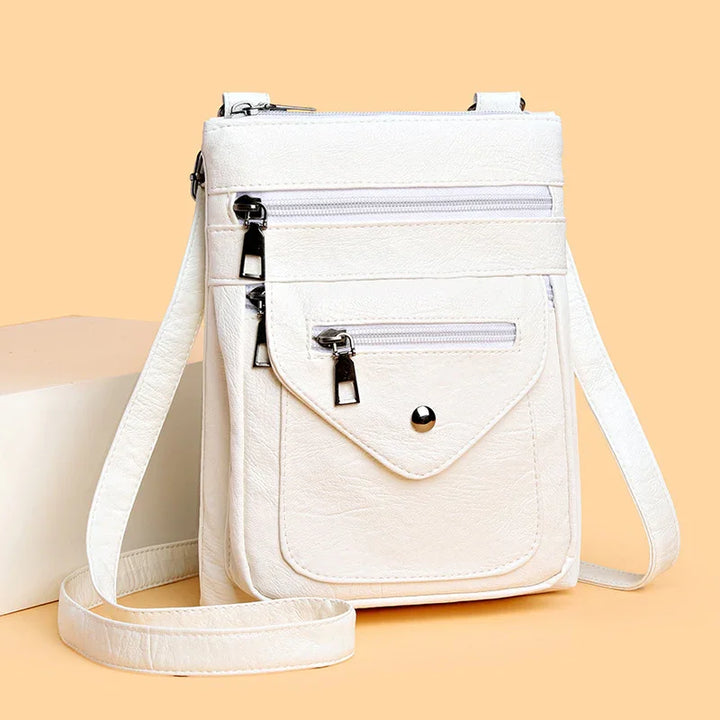 White crossbody bag with multiple zippered pockets and adjustable shoulder strap on peach background