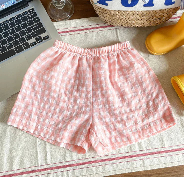 Pink and white gingham textured shorts on a table with laptop, wicker basket, and yellow rain boots