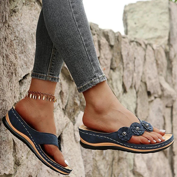 Woman wearing navy blue wedge sandals with floral details and ankle chain, standing by stone wall