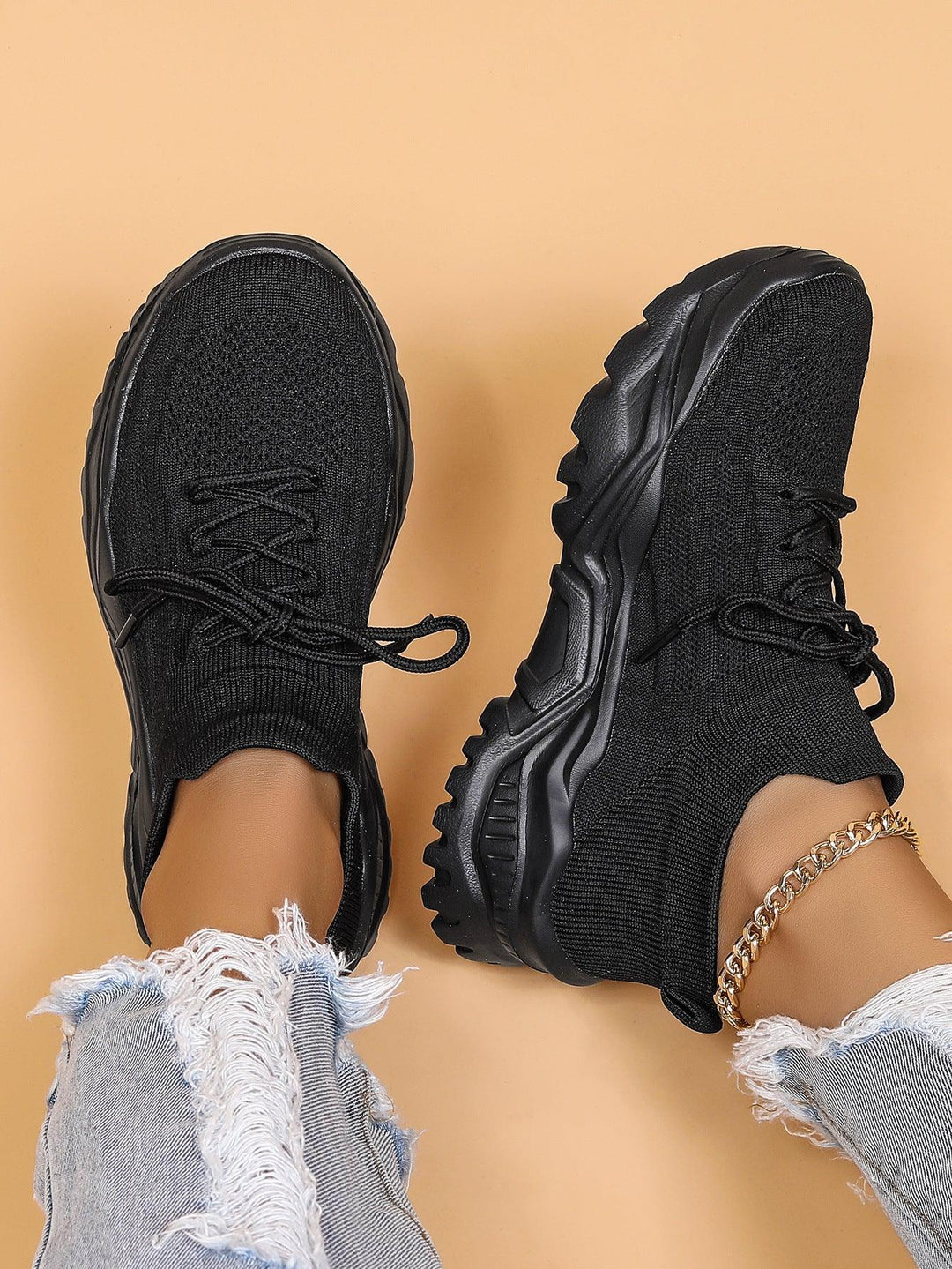 Black knitted sock sneakers with thick textured soles worn with ripped light blue jeans and gold anklet on peach background