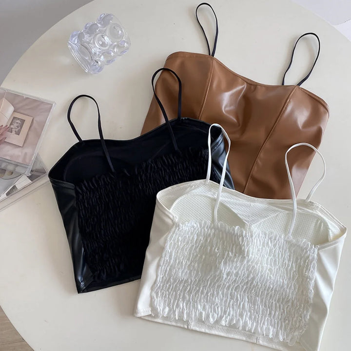 Three leather crop tops with spaghetti straps in brown, black, and white on a white table with a glass and photos.