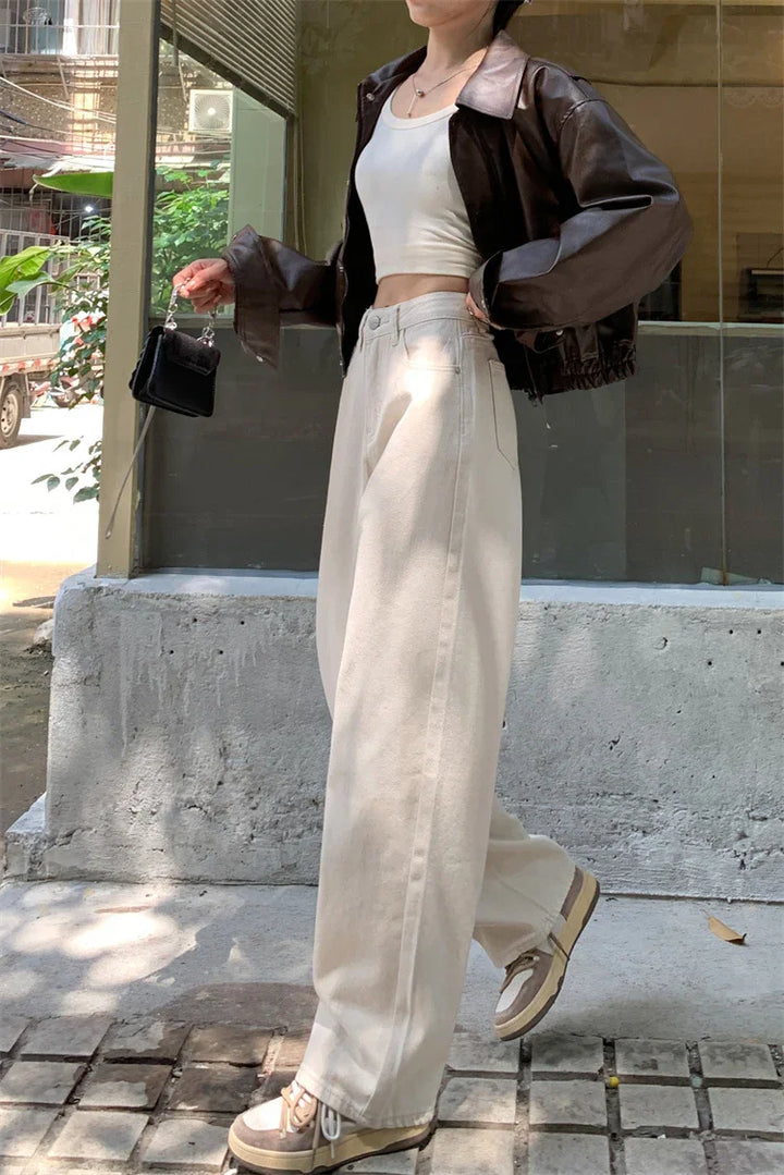 woman wearing cropped brown leather jacket, white crop top, wide-leg beige pants, and beige sneakers holding small black purse