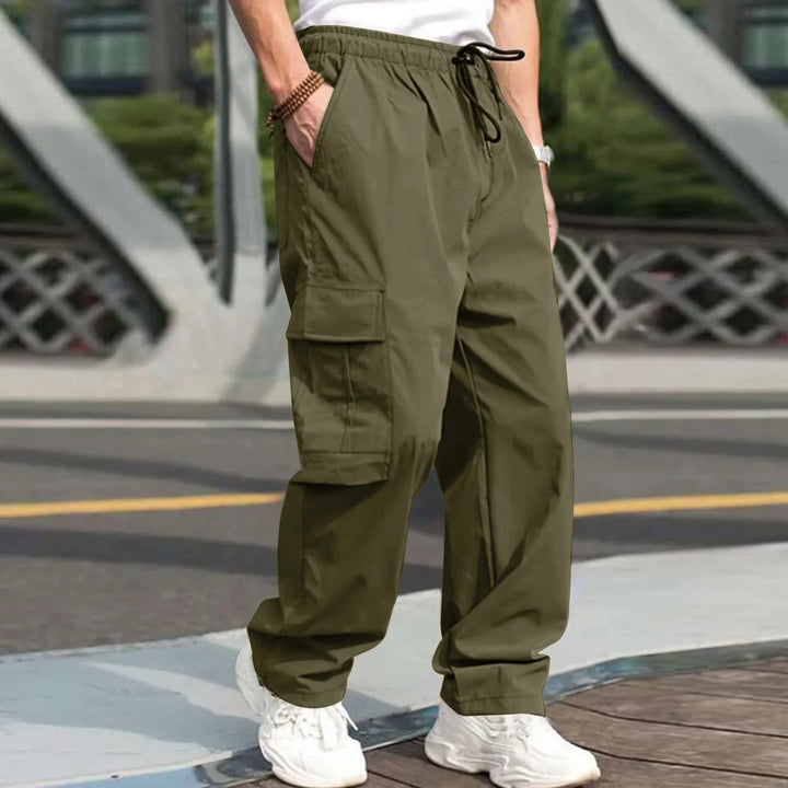 Man wearing olive green cargo pants and white sneakers standing outdoors near bridge railing