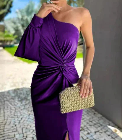 Woman wearing elegant one-shoulder purple dress with knot detail and holding a gold clutch outdoors
