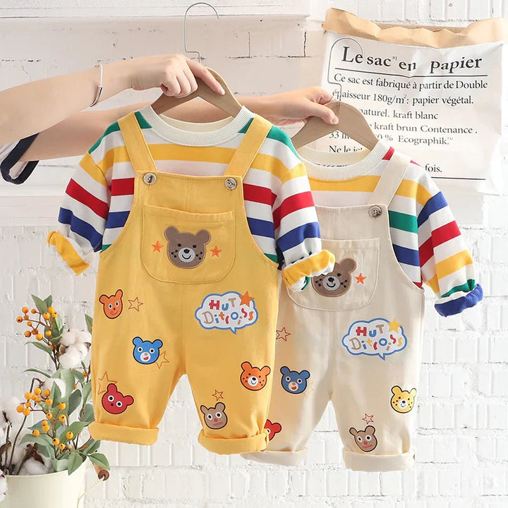 Two toddler overalls with colorful bear patches and striped long-sleeve shirts hanging on wooden hangers