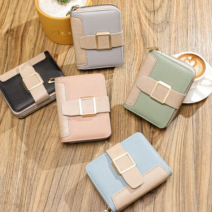 Five compact leather wallets in black, gray, pink, green, and blue on wooden surface with cappuccino and potted plant