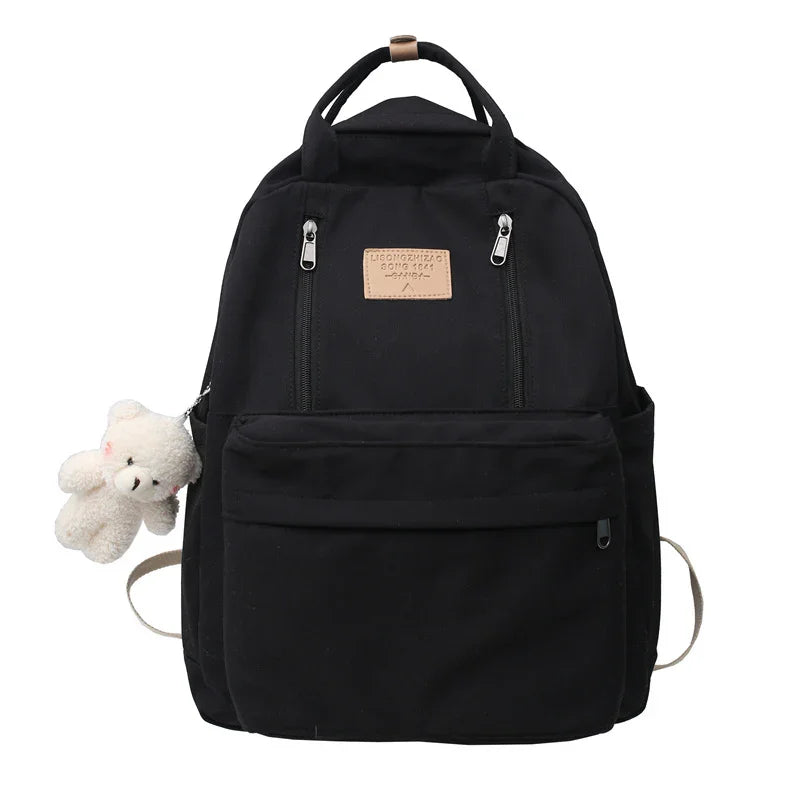 Black canvas backpack with front zipper pockets and a small white teddy bear keychain attached