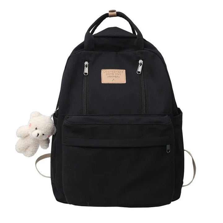 Black canvas backpack with front zipper pockets and a small white teddy bear keychain attached