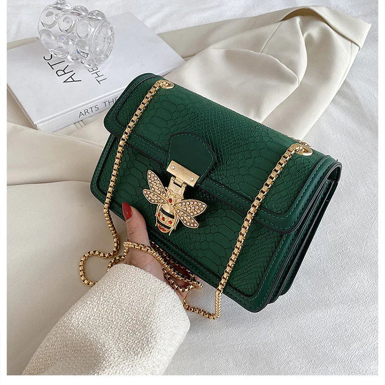 Hand holding a green textured leather handbag with a gold bee clasp and chain strap on white fabric