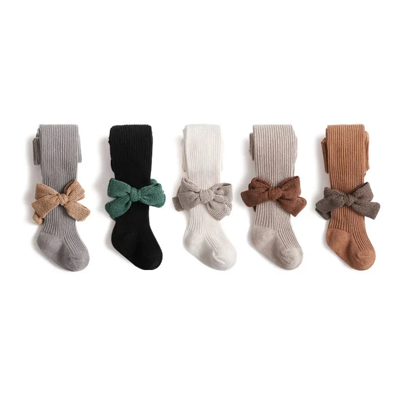 Five pairs of ribbed baby tights in gray, black, white, beige, and brown with matching knit bows on a white background