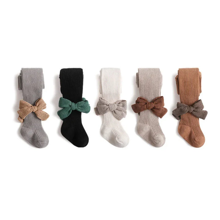 Five pairs of ribbed baby tights in gray, black, white, beige, and brown with matching knit bows on a white background