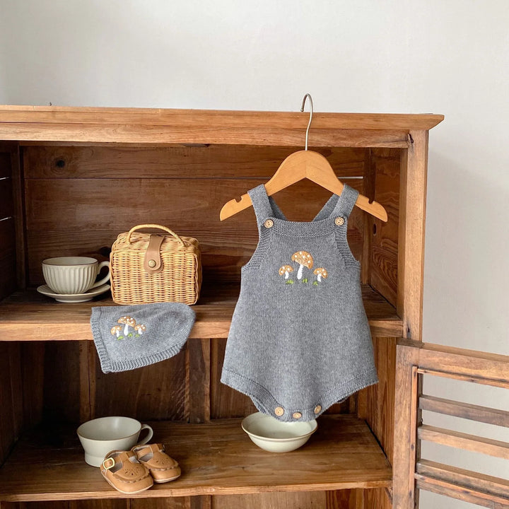 Gray knitted baby romper and matching hat with mushroom embroidery displayed on wooden shelves