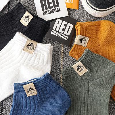 Assorted ribbed cotton socks in black, white, blue, green, and mustard yellow with labels on a carpet next to red charcoal boxes