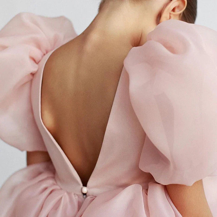 Back view of a woman wearing a light pink dress with sheer puff sleeves and low V-cut back detail