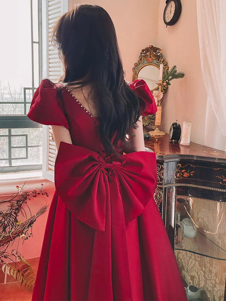 Woman in elegant red dress with large bow back detail standing by window near antique dresser
