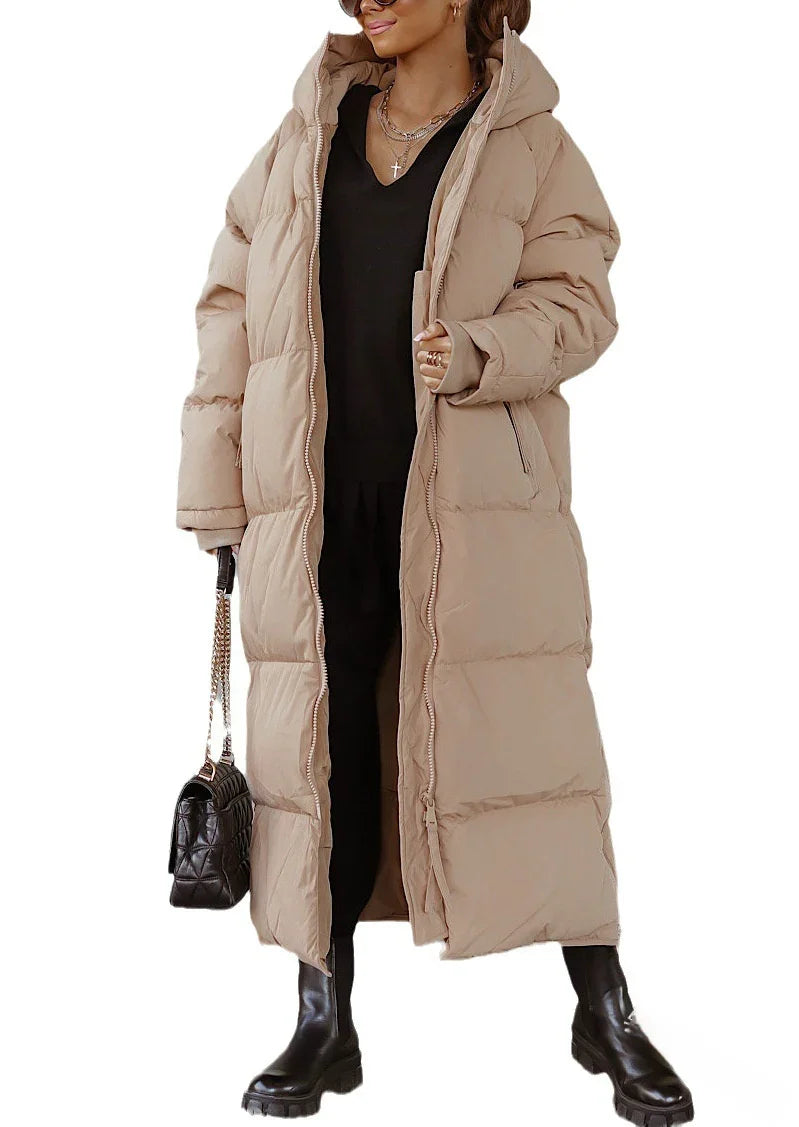 Woman wearing long beige puffer coat with hood, black outfit, black boots, and holding black handbag
