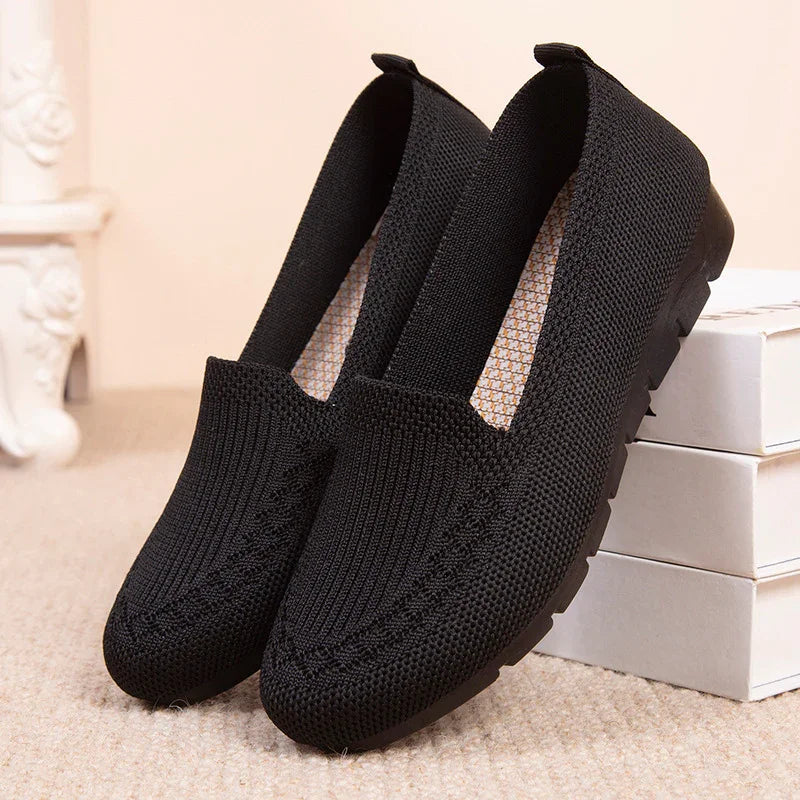 Pair of black knitted slip-on casual shoes on beige carpet with white boxes in background