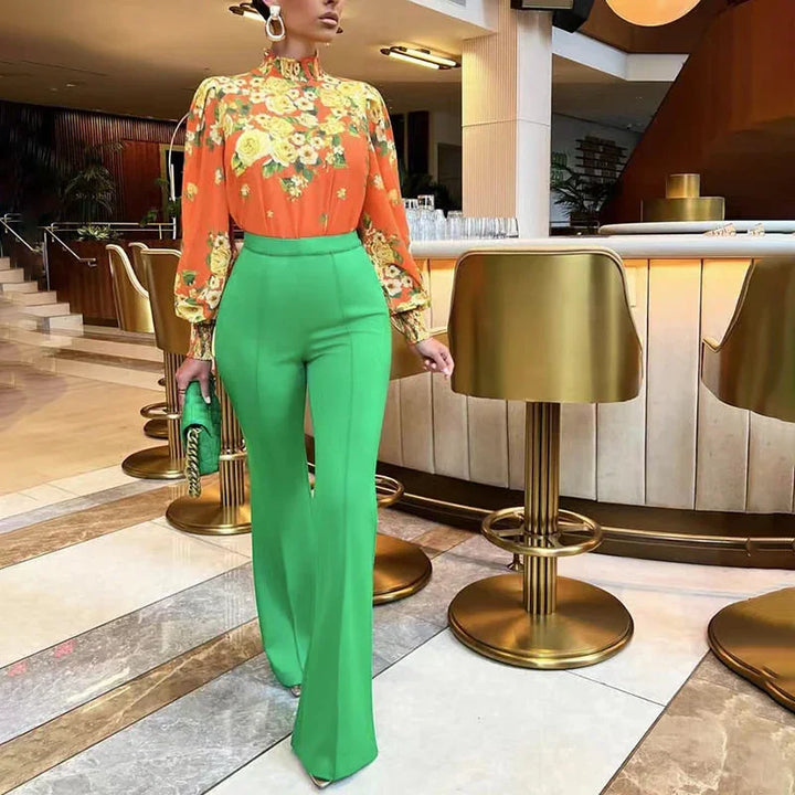 Woman wearing orange floral blouse and high-waisted green pants holding green clutch in modern bar