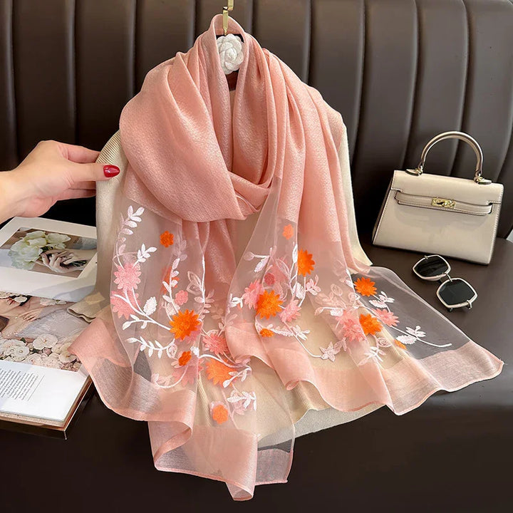 Pink sheer scarf with orange and white floral embroidery draped on beige chair with cream handbag and sunglasses
